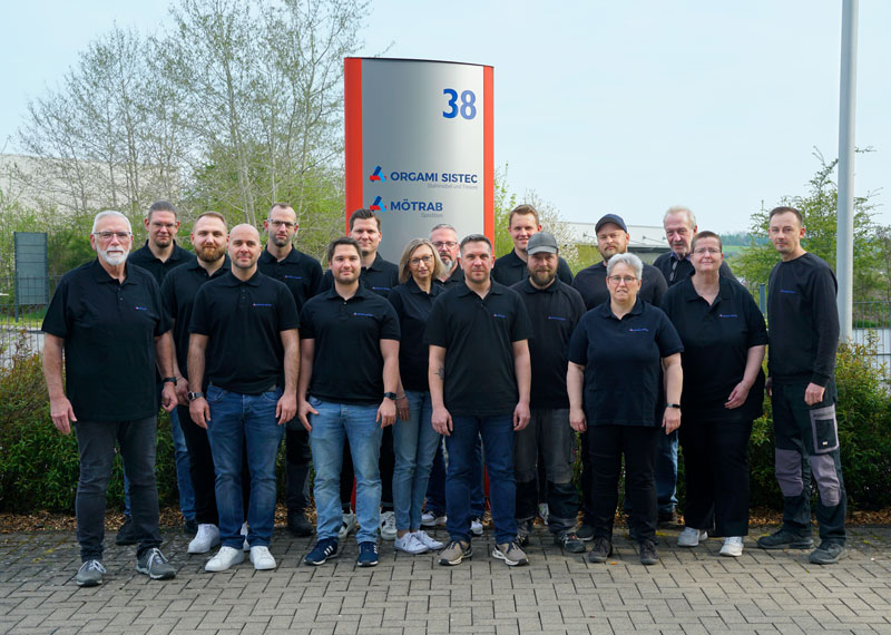 Eine Gruppe von 17 Mitarbeitern steht draußen vor einer Hecke mit Zaun. Im Hintergrund befindet sich ein Schild mit der Nummer 38 und den Firmennamen „Orgami Sistec“ und „Mötrab“. Zudem sind einige Bäume zu sehen, und der Himmel ist blau-grau.