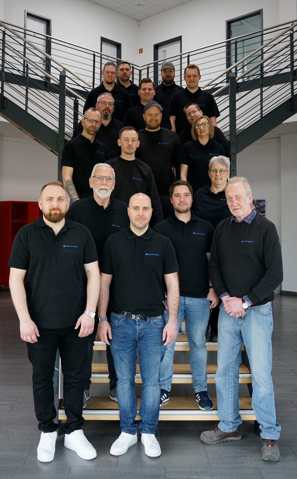 Ein Bild von 17 Mitarbeitern auf einer Treppe in der Eingangshalle eines Bürogebäudes. Alle tragen ein schwarzes T-Shirt mit dem Firmenlogo auf der rechten Brustseite. Die Wand im Hintergrund ist weiß.