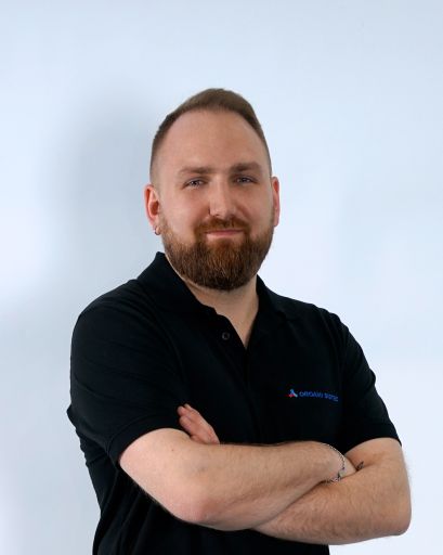 Bild von einem Mann mit Bart. Er trägt ein schwarzes T-Shirt mit der Aufschrift „Orgami Sistec“ + Logo. Stehend vor einem weißen Hintergrund.