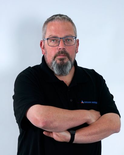 Bild von einem Mann mit Bart und Brille. Er trägt ein schwarzes T-Shirt mit der Aufschrift „Orgami Sistec“ + Logo. Stehend vor einem weißen Hintergrund.