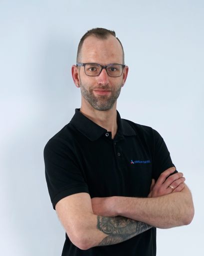 Bild von einem Mann mit Bart und Brille. Er trägt ein schwarzes T-Shirt mit der Aufschrift „Orgami Sistec“ + Logo. Stehend vor einem weißen Hintergrund.