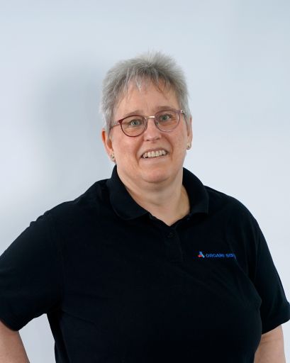 Bild von einer Frau mit kurzen Haaren und einer Brille. Sie trägt trägt ein schwarzes T-Shirt mit der Aufschrift „Orgami Sistec“ + Logo. Stehend vor einem weißen Hintergrund.
