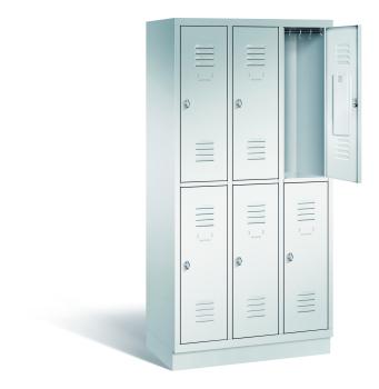 Ein lichtgrauer Garderobenschrank mit drei Abteilen, sechs Fächern sowie integrierten Kleiderstangen mit Kleiderhaken, dessen obere rechte Tür geöffnet ist. Er steht auf einem Stahlblechsockel vor weißem Hintergrund.