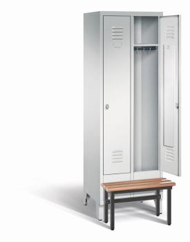 Ein lichtgrauer Garderobenschrank mit zwei Abteilen, zwei Fächern sowie integrierten Kleiderstangen mit Kleiderhaken, dessen rechte Tür geöffnet ist. Er steht auf Stahlblechfüßen und hat eine vorgebaute Sitzbank vor weißem Hintergrund.