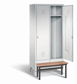 Ein lichtgrauer Garderobenschrank mit zwei Abteilen, zwei Fächern sowie integrierten Kleiderstangen mit Kleiderhaken, dessen rechte Tür geöffnet ist. Er steht auf Stahlblechfüßen und hat eine vorgebaute Sitzbank vor weißem Hintergrund.