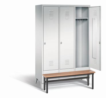 Ein lichtgrauer Garderobenschrank mit drei Abteilen, drei Fächern sowie integrierten Kleiderstangen mit Kleiderhaken, dessen rechte Tür geöffnet ist. Er steht auf Stahlblechfüßen und hat eine vorgebaute Sitzbank vor weißem Hintergrund.