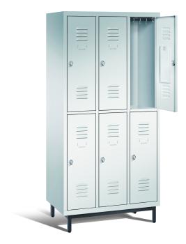 Ein lichtgrauer Garderobenschrank mit drei Abteilen, sechs Fächern sowie integrierten Kleiderstangen mit Kleiderhaken, dessen obere rechte Tür geöffnet ist. Er steht auf Stahlrohrfüßen vor weißem Hintergrund.