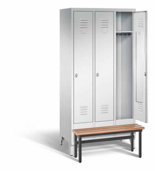 Ein lichtgrauer Garderobenschrank mit drei Abteilen, drei Fächern sowie integrierten Kleiderstangen mit Kleiderhaken, dessen rechte Tür geöffnet ist. Er steht auf Stahlblechfüßen und hat eine vorgebaute Sitzbank vor weißem Hintergrund.