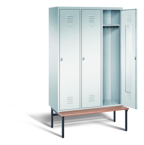 Ein lichtgrauer Garderobenschrank mit drei Abteilen, drei Fächern sowie integrierten Kleiderstangen mit Kleiderhaken, dessen rechte Tür geöffnet ist. Er steht auf einer unter- und vorgebauten Sitzbank vor weißem Hintergrund.