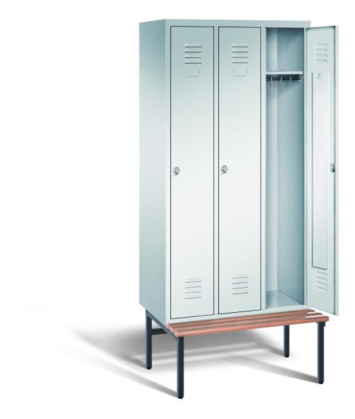 Ein lichtgrauer Garderobenschrank mit drei Abteilen, drei Fächern sowie integrierten Kleiderstangen mit Kleiderhaken, dessen rechte Tür geöffnet ist. Er steht auf einer unter- und vorgebauten Sitzbank vor weißem Hintergrund.