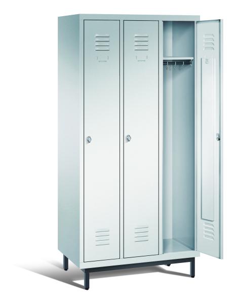 Ein lichtgrauer Garderobenschrank mit drei Abteilen, drei Fächern sowie integrierten Kleiderstangen mit Kleiderhaken, dessen rechte Tür geöffnet ist. Er steht auf Stahlrohrfüßen vor weißem Hintergrund.